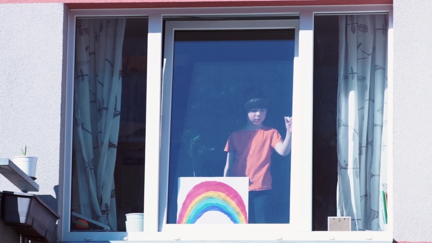 Young schoolboy puts a homemade rainbow paint drawing on window of his house. Self isolation and positivity amid  during covid-19 quarantine. “Chase the rainbow” trend. Cz, Kladno, G.Selnera 08.4.2020