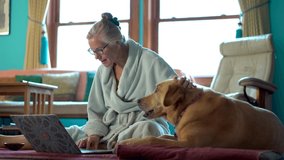 Work from home mature woman in bathrobe on the floor with laptop petting and talking to dog and feeding pet on the floor.