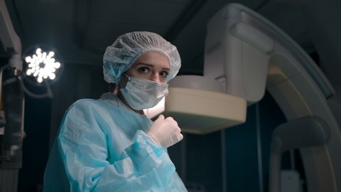 Low angle of young woman in surgeon uniform and gloves taking off mask and smiling after successful operation in modern operating room.