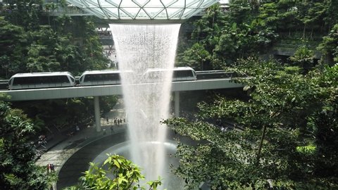 Singapore - Aug 8, 2019: skytrain on suspended railway bridge transporting people to terminal Changi International Airport. On background the Rain Vortex, indoor waterfall inside Jewel Airport.