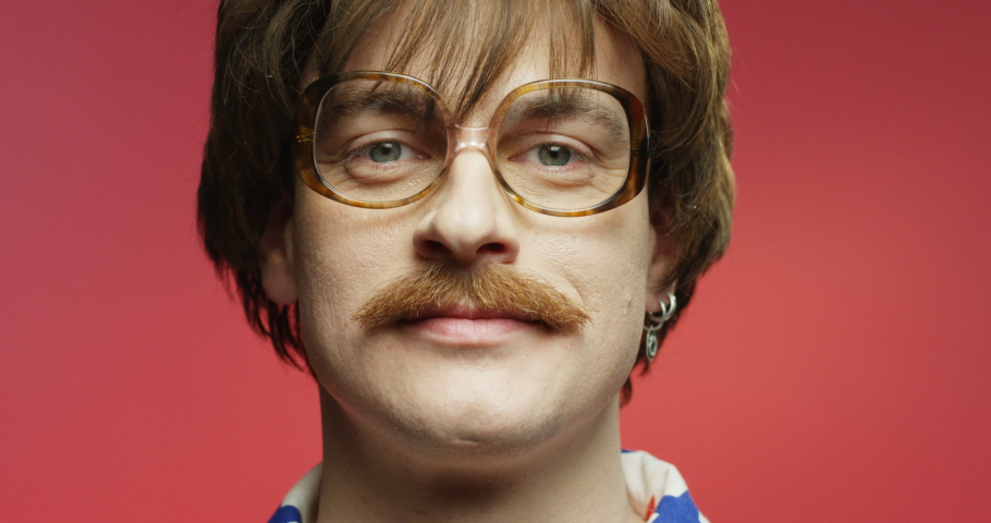 Funny retro style dark-haired Caucasian man with mustache and eyeglasses smiling to camera. Close up portrait of happy male over pink background. Vintage stylish model with smile on face. 70s concept