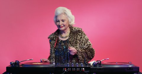 Retro stylish Caucasian female pensioner in fancy outfit of 70s 80s playing music at DJ equipment over pink wall at party. Vintage concept of old woman working as dj disco.