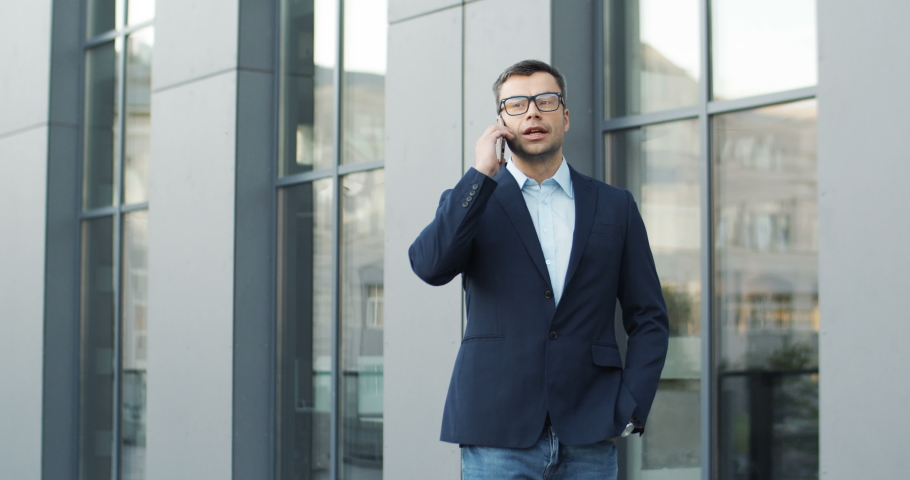 Caucasian handsome businessman in glases standing outdoors and talking on his smartphone. Attractive man speaking on mobile phone while chatting at street. Male using cellphone.