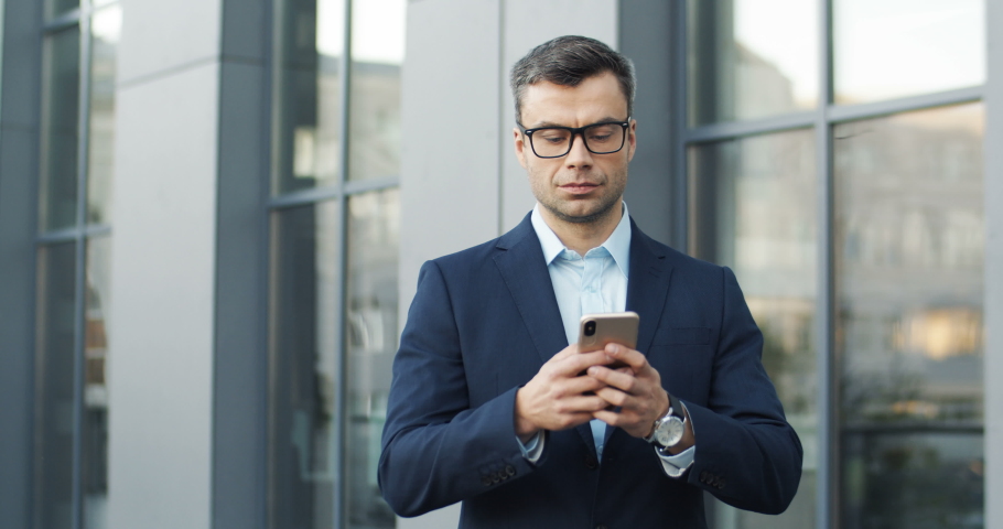 Caucasian handsome businessman in glases standing outdoors and holding his smartphone. Attractive man tapping ad texting message on mobile phone while chatting at street. Male using cellphone.