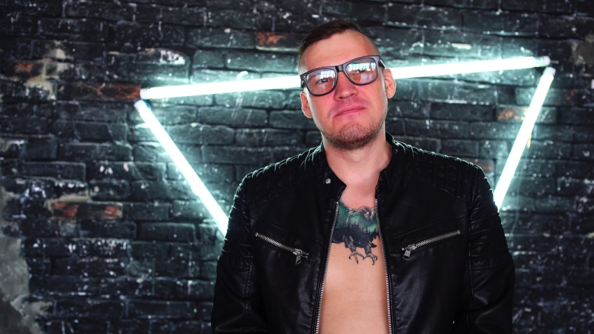 Young man with glasses scratching his chin thoughtfully on background shabby brick wall with glowing lamps in form of triangle.