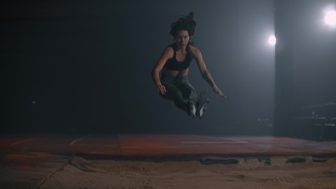 Running woman athletics at the stadium on a treadmill performs a triple jump in length and flies into the sand landing on her feet. Slow motion