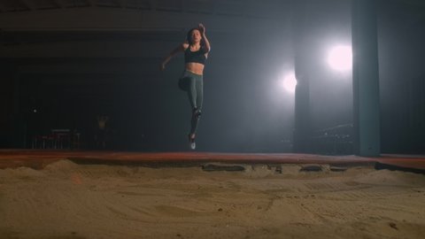 A sporty woman runs up in a stadium and performs a long jump in slow motion in a stadium on a dark background