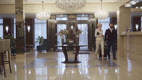 Extreme wide shot of Caucasian man and woman with travel bag entering luxurious hotel vestibule and approaching to reception desk. Rich couple checking in at high-class accommodations. Tourism, luxury