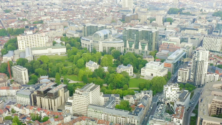 Brussels, Belgium. The European Parliament complex at Espace Leopold sits within a lush green park, surrounded by the dense urban fabric of the European Quarter. The scene centers on the distinctive postmodern architecture of the parliament buildings, with curved glass facades and a prominent oval structure rising among mature trees and landscaped grounds. The complex occupies a former industrial site that was transformed into one of the primary seats of European governance. The surrounding area blends modern institutional architecture with historic residential buildings, all viewed from a high aerial perspective that reveals the full scale of the compound and its integration into the city. Bright daylight illuminates the scene, with clear skies enhancing the contrast between the deep green of the park and the pale stone and reflective glass of the buildings. The dolly zoom effect creates a dynamic sense of depth, drawing the viewer