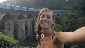 Young woman taking selfie while train passing on old railway bridge in the middle of tea plantations. Gril selfie video in Sri Lanka 