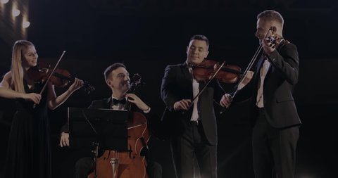 Cheerful men and woman from quartet of classical music having a concert. Male and female musicians performing their music on dark stage in spotlight. Orchestra playing on violins and cello.