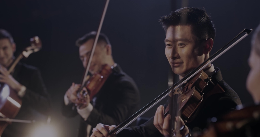 Close up of smiled hapy male young Asian violinist playing on violin in classical orchestra with Caucasian colleague at concert. Cheerful musician performing on his instrument.