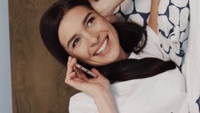 Vertical video of beautiful happy girl talking on cellphone while lying in bed at home.Close up portrait of pretty Caucasian cheerful young woman smiling and speaking phone in bedroom. Rest concept