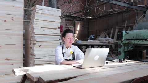 woman working as carpenter in her own woodshop. She using a laptop and writes notes while being  in her workspace. Small business concept.: film stockowy