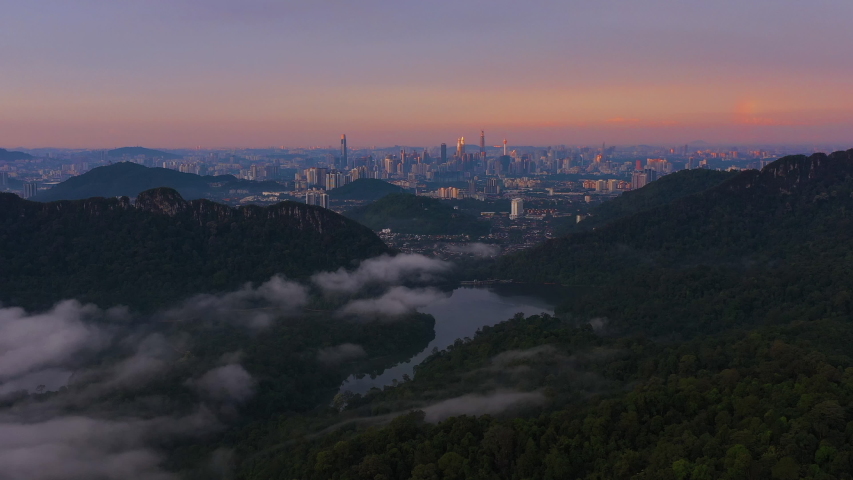 4K Aerial view flying over lake and lush green tropical foggy rain forest with city skyline in background. Rolling clouds in colourful early morning sunrise in Kuala Lumpur. (Noise is visible)