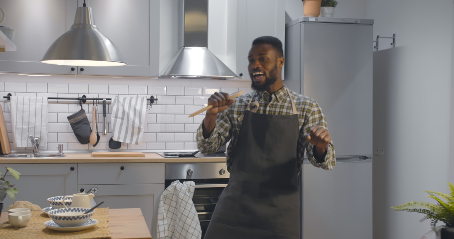 Portrait of cheerful young afro-american man in apron dancing and singing using wooden spoon as microphone cooking dinner in modern kitchen. Funny guy preparing meal Royalty-Free Stock Footage #1050217621