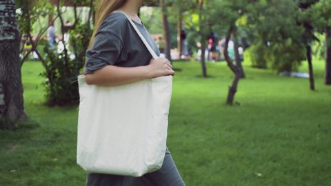 Side view of Young girl holds white textile eco bag and walks in the summer park