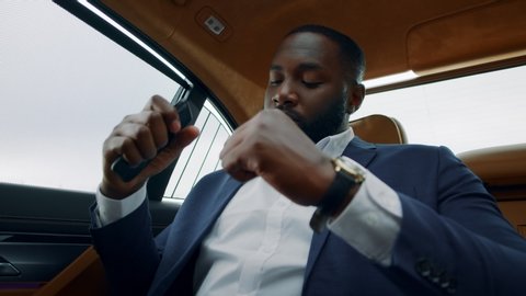 Portrait of successful afro man dancing in luxury car. Happy african businessman listening music on mobile phone at backseat. Stylish black business man dancing with smartphone in hand.