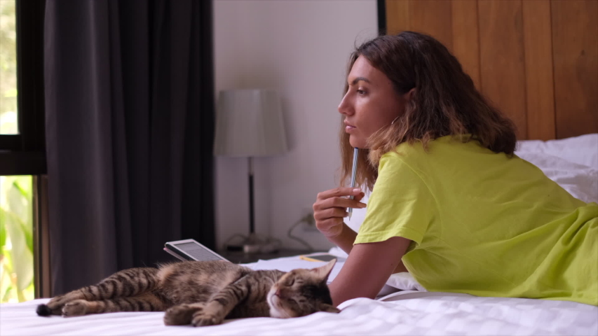 The girl lies with a pensive look on the bed and thinks about something. Woman lies with a cat in the bedroom and meditates with a pen in her hand and an e-book