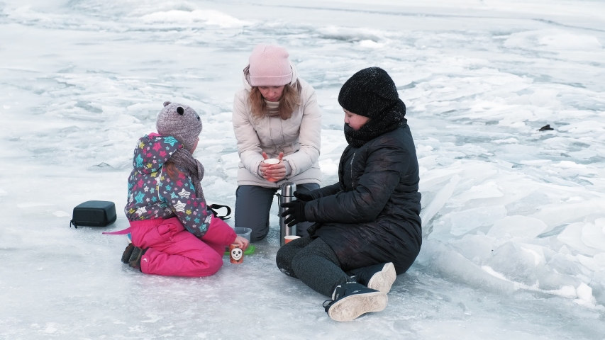 Family picnic on a frozen river. Mom with daughters drinks tea on a winter river.