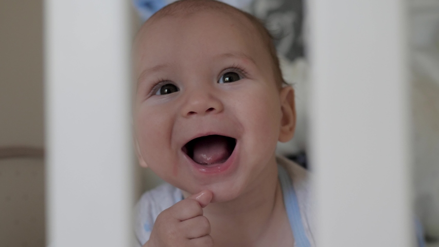 A nice little baby is smiling cheerfully through the sides of the crib. Portrait of a playful and energetic child close-up, he sucking a finger. Royalty-Free Stock Footage #1050275470