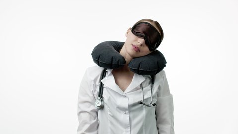 Beautiful sexy female doctor in medical uniform with stethoscope puts on sleeping mask and portable travel neck pillow, playfully winks and fall asleep. Sleeping at work, taking nap, resting concept