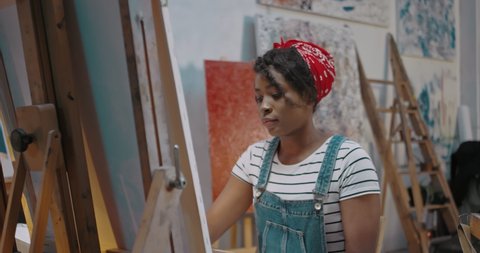 Young African American stylish female artist painting impressionism picture on canvas and creating artwork in art studio. Pretty girl student at fine art course. Beautiful woman studying at academy.