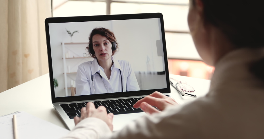 Videoconferencing doctor concept. Female therapist wears headset providing remote medical assistance in virtual conference chat consulting patient online by webcam on laptop screen. Over shoulder view Royalty-Free Stock Footage #1050311641
