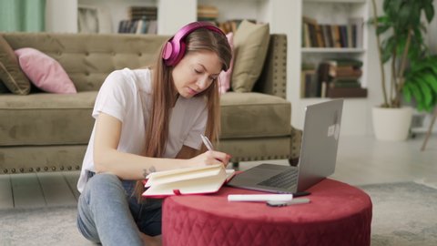 Girl student studying at home reading notes in exercise book using computer for online self education doing homework.Young woman with blond hair in headphones reading a book and taking notes. 4k