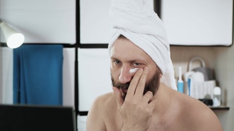 Man applies cream around eyes. Travesty, attractive man in bathroom with towel on his head applies cream to his face in eye area. Close-up