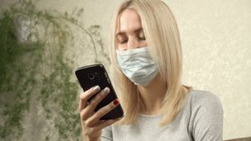 Woman in a medical protective face mask at home quarantine. Girl using mobile phone, video call. Pandemic mood. Woman speaking online, internet communication. Self-isolation during epidemic.