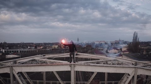 Aerial view of young man in hoodie and balaclava standing on the top of the frame construction of an old steel frame bridge with red burning signal flare