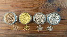 Cereal grains in glass jars on wooden background 4K video. Collection of different groats top view barley, oats, millet and wheat. Wholegrain foods high fiber content flat lay.Healthy diet ingredients