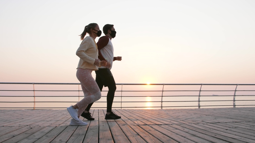 Young fit couple in protective masks running outdoors near the sea during beautiful sunrise, slow motion, low angle view