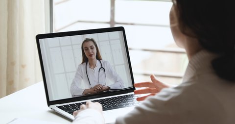Young woman patient talking with online female doctor therapist in webcam video call app on laptop screen. Remote telemedicine healthcare medical consultation concept. Over shoulder close up view