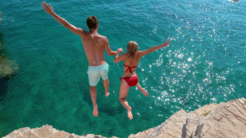 SLOW MOTION: Carefree tourists hold hands while jumping into the refreshing sea during a relaxing summer vacation in Croatia. Woman and her boyfriend dive off a cliff and into the deep blue ocean. Royalty-Free Stock Footage #1050377563