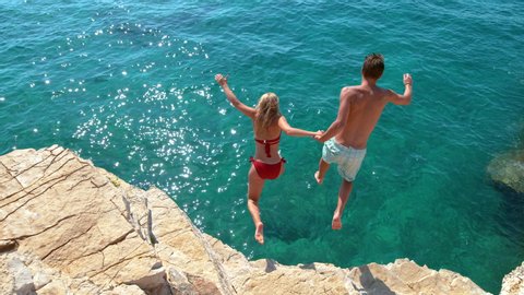 SLOW MOTION: Joyful tourist couple decides to jump off a rocky cliff and dive into sea. Unrecognizable man holds his girlfriend's hand while diving into the deep blue sea on a sunny summer day.