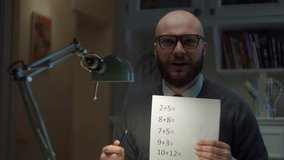 Bearded 30s man in glasses showing sheet of paper with simple mathematical equations looking at camera at home. Online education process. Webcam view of teacher talking. 