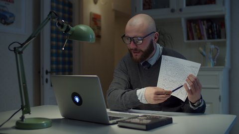 Bearded 30s man in glasses showing sheet of paper with graphs for students looking at laptop at home. Online education process. Side view of teacher talking. 