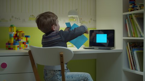 Preschooler boy making online art homework. Kid cutting blue square shape with scissors sitting at the desk in children room at home.