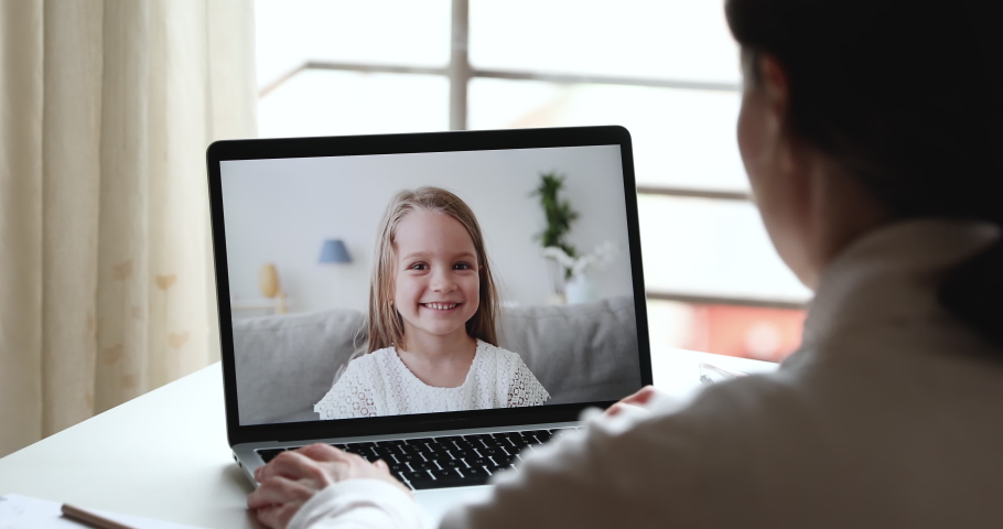 Female tutor video calling teaching cute preschool kid girl using remote learning app talking with happy child in chat on laptop screen. Home conference children education. Over shoulder closeup view Royalty-Free Stock Footage #1050385018