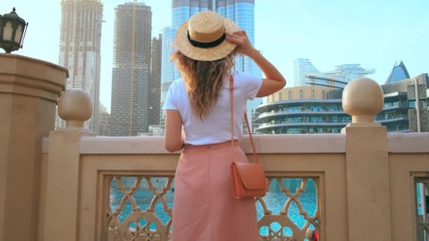 happy cute woman tourist in straw hat enjoys vacation back view. modern skyscrapers city center Dubai. United Arab Emirates UAE 2020. looks up to landmark hight building. blue sky water. Go Everywhere