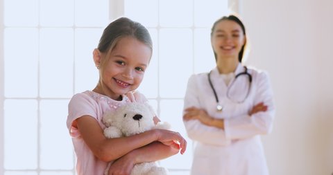 Happy small cute kid girl embracing fluffy teddy bear toy looking at camera with smiling young female pediatrician doctor in white uniform with stethoscope on background, children healthcare concept.