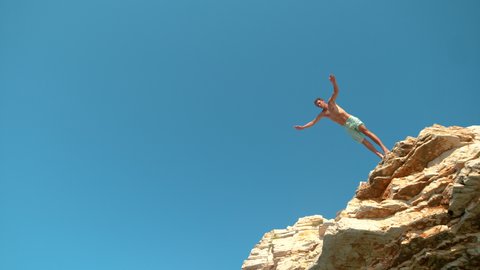 SLOW MOTION, BOTTOM UP: Joyful Caucasian man on summer vacation rock diving on a sunny day. Athletic male tourist jumps off a rocky cliff to cool off in the refreshing ocean from the summer heat.