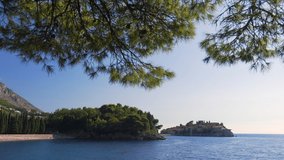 Tourist destination-Montenegro. Video of the place Sveti Stefan island in Budva. View of the island and the beach from the shore through the branches of pine trees. Bright sunny day