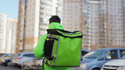 Panning shot of young male food courier wearing protective mask while walking down city street with insulated backpack delivering order