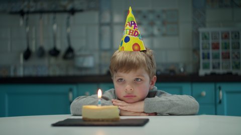 Young sad preschooler boy in birthday hat looking to candle on piece of cake. Lonely upset birthday kid sitting at home on kitchen. 