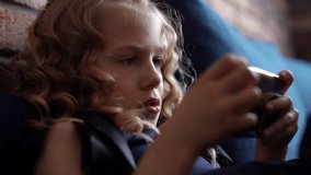 Close-up of face of focused curly beautiful little girl playing video games on smartphone while sitting on couch at home in cozy living room. Shotting in slow motion.