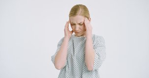 Camera zooming in at face of blonde Caucasian ill woman on white wall background touching with fingers her forehead. Sick weak female patient having migraine headache. Disease symptom.