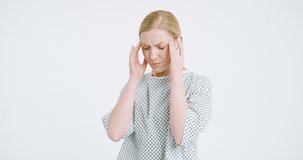 Camera zooming in at young Caucasian sick woman in hospital night dress standing on white background and holding with fingers her forehead. Ill female patient having migraine headache. Virus symptoms.